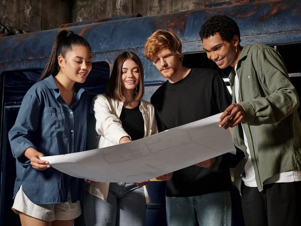 Escape room website design: group of smiling friends studying a large map for clues together in a dim industrial themed room