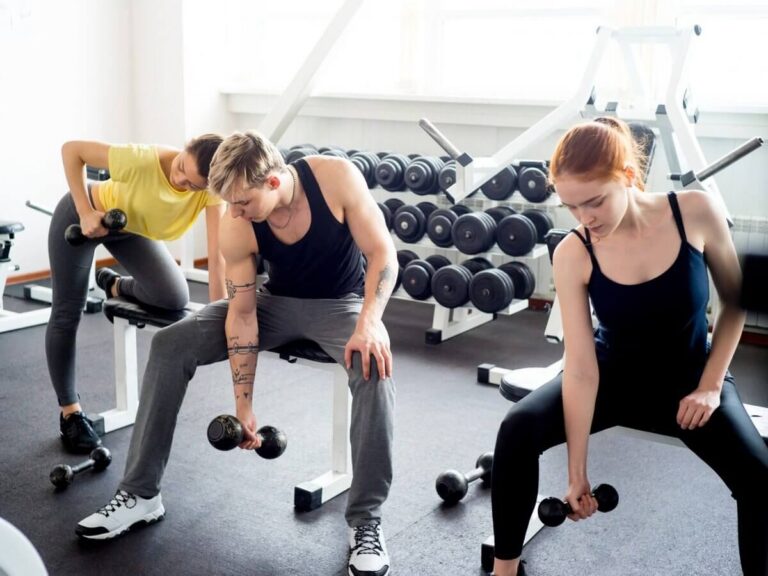 Gym website design: gym members training with dumbbells on benches in a bright fitness studio with a full weight rack