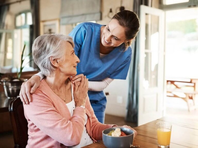 Home care agency website design: home caregiver in blue scrubs smiling with an older woman at her breakfast table