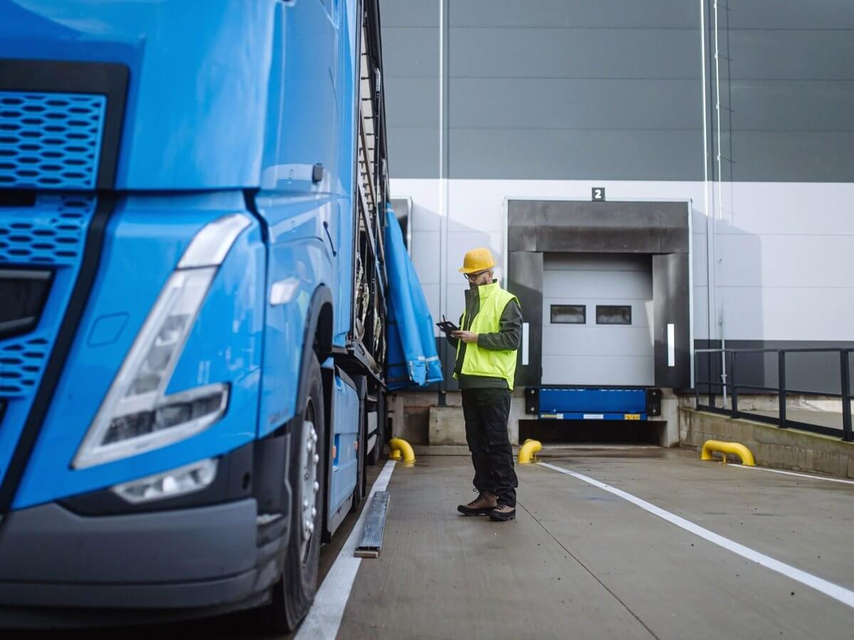 Logistics company website design: logistics worker in a hi-vis vest checking a tablet beside a semi truck at a warehouse loading dock