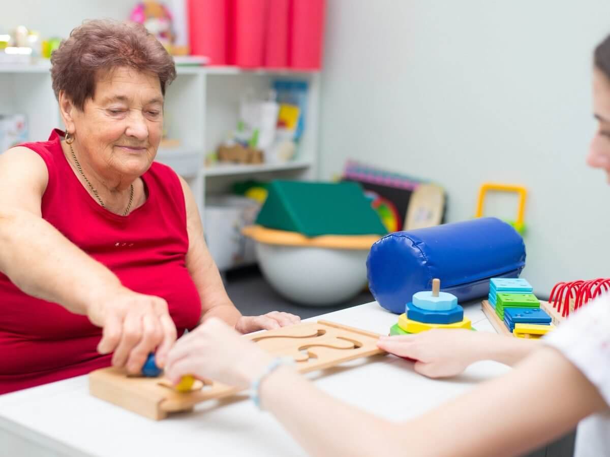 Occupational therapy practice website design: older client working a wooden fine motor board with an occupational therapist guiding her hands in a bright clinic